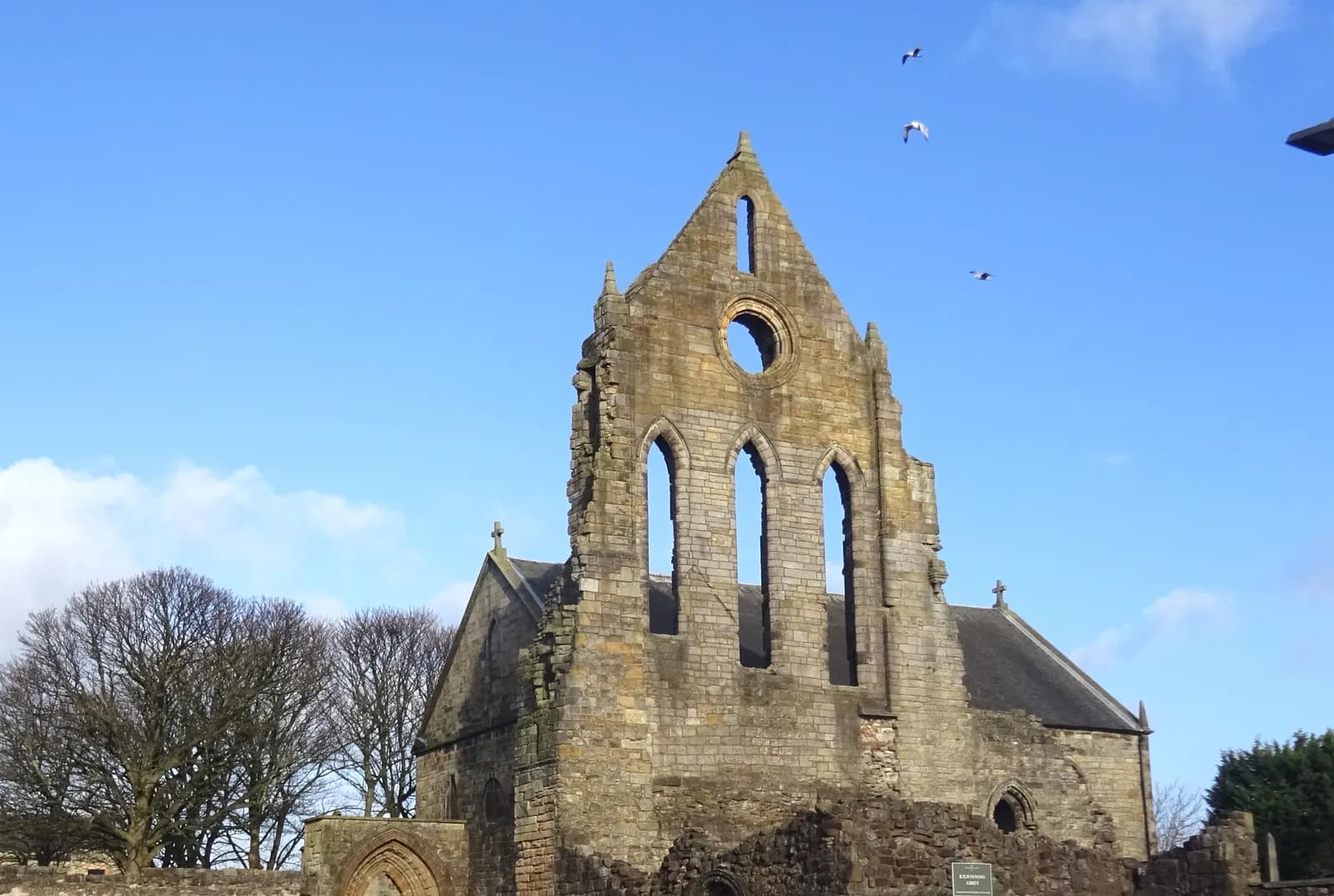 The ruined gable of Kilwinning Abbey against a blue sky, with the Abbey Church behind