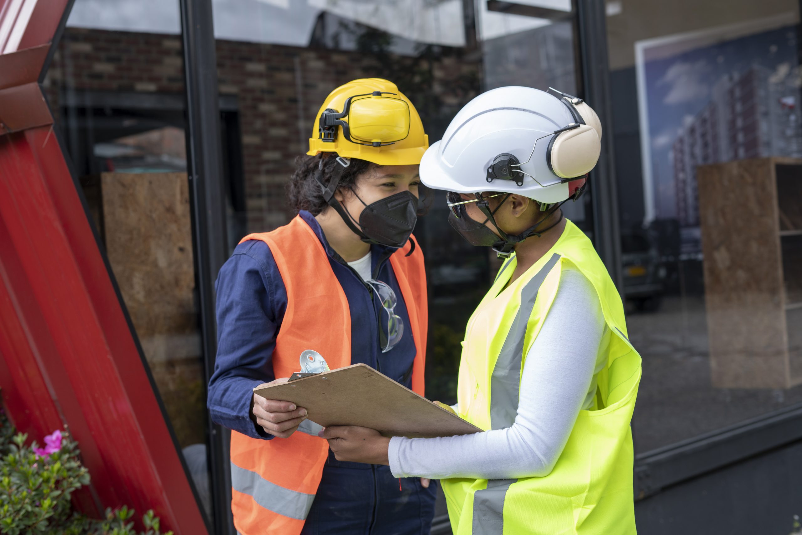 Two uniformed Afro-skinned and Afro-haired Latina women with reflective vests and protective helmets with face masks in the middle of the building that they lead by observing the plans